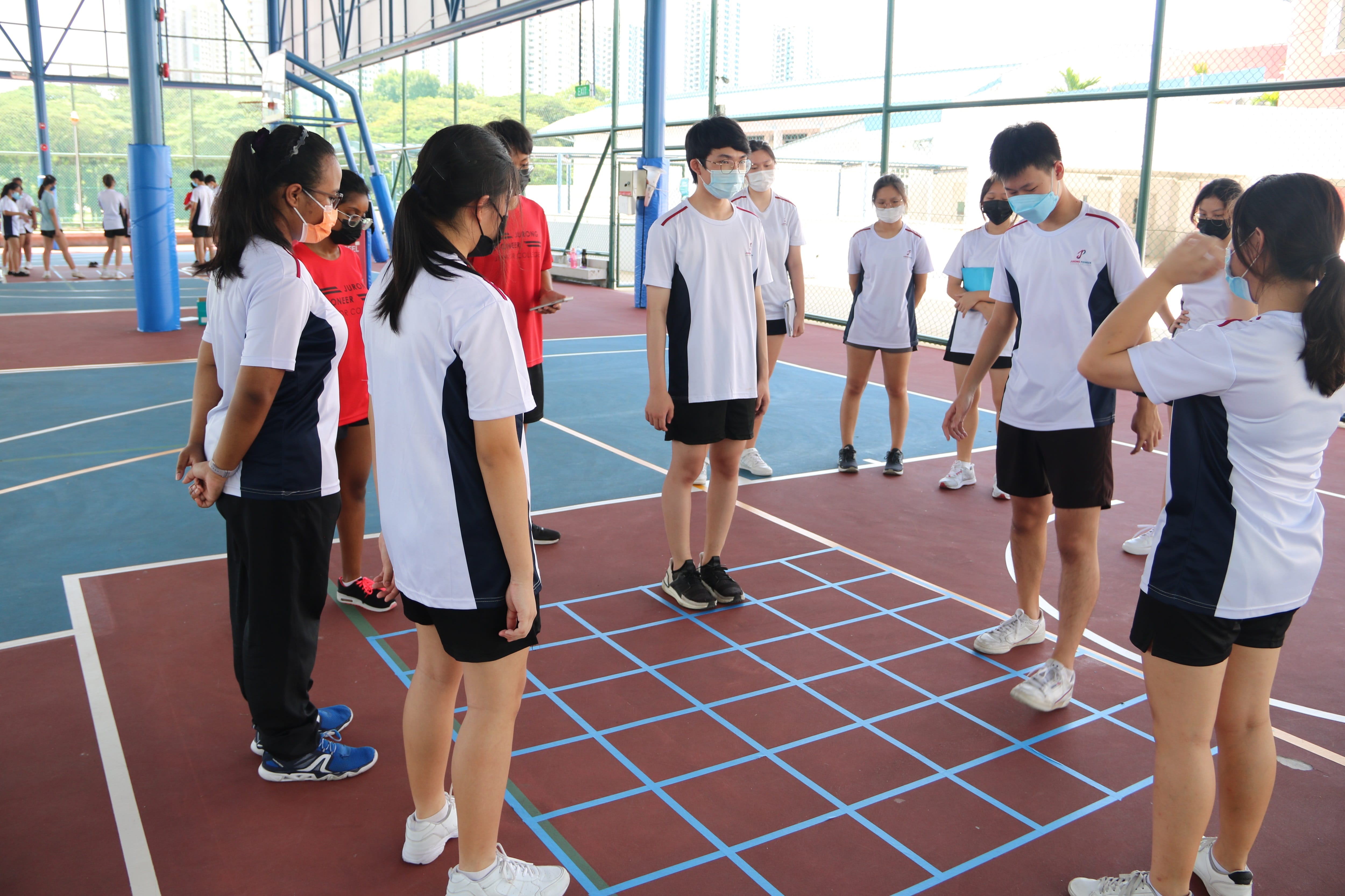 Students in white uniforms and face masks stand in a circle on an indoor sports court with blue grid lines.